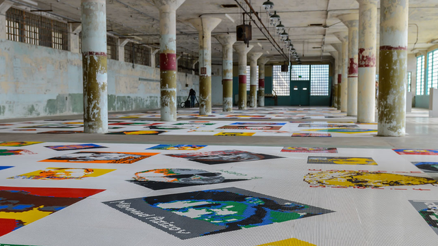 Lego brick pieces are arranged to create portraits along a floor inside a large concrete room with columns