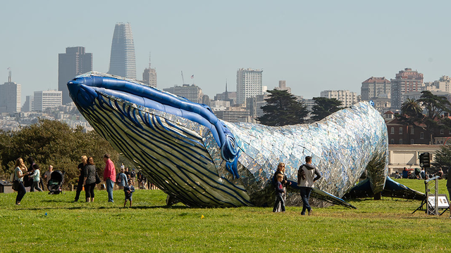 People wander around a huge sculpture of a blue whale, with the San Francisco skyline in the distance