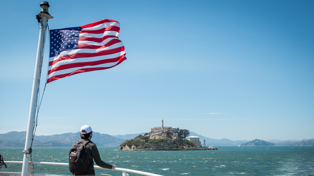 A park visitor looks toward Alcatraz Island from the ferry. An American flag waves in the breeze as they cruise over bay waters.