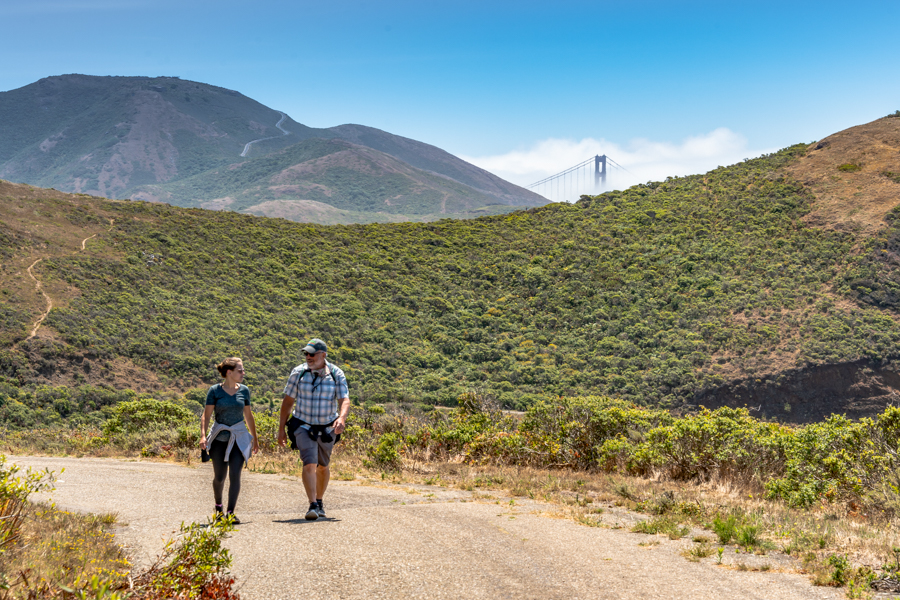 A couple hiking a dirt trail in the Marin Headlands. A Golden Gate Bridge tower can be seen peeking through the green mountain range in the background.