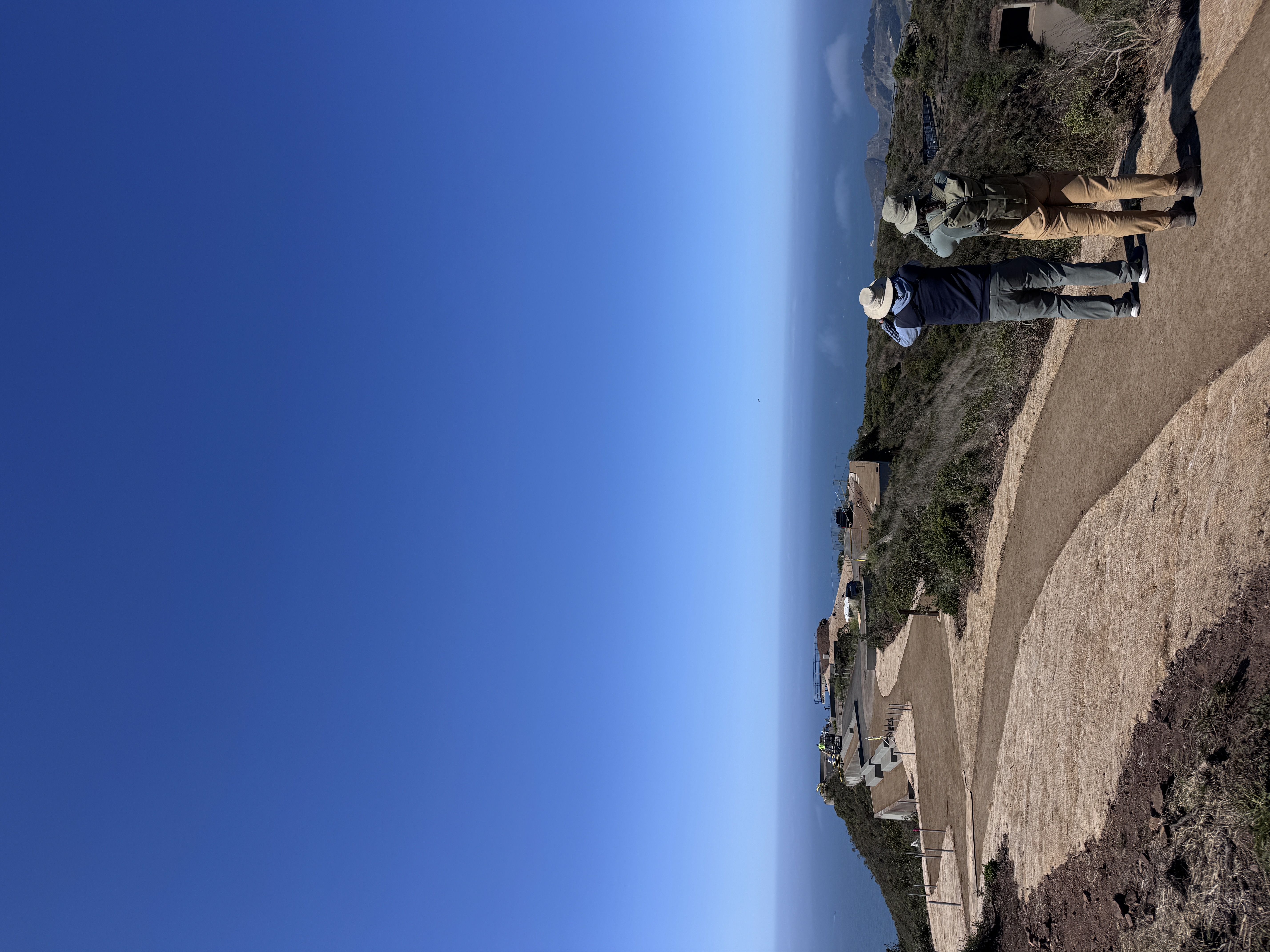 To volunteers look through binoculars on botton right, standing on a hilltop against a blue sky.
