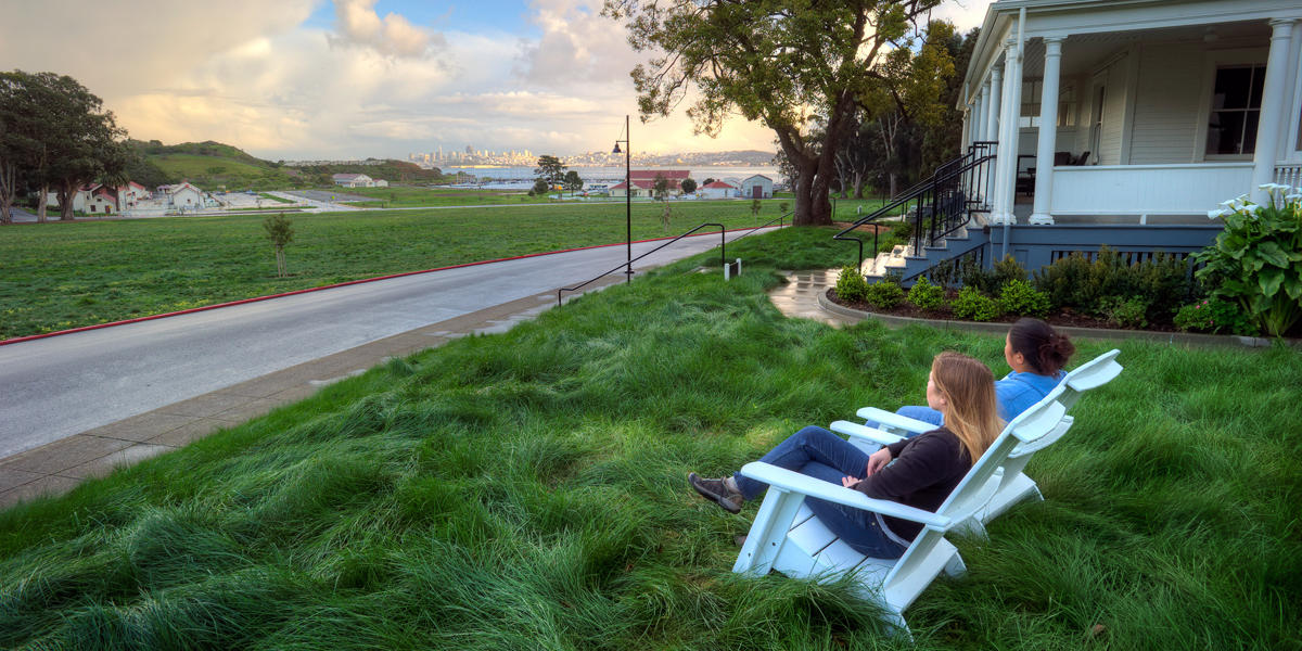 Two people sit in chairs and look out over a grassy field at the San Francisco skyline