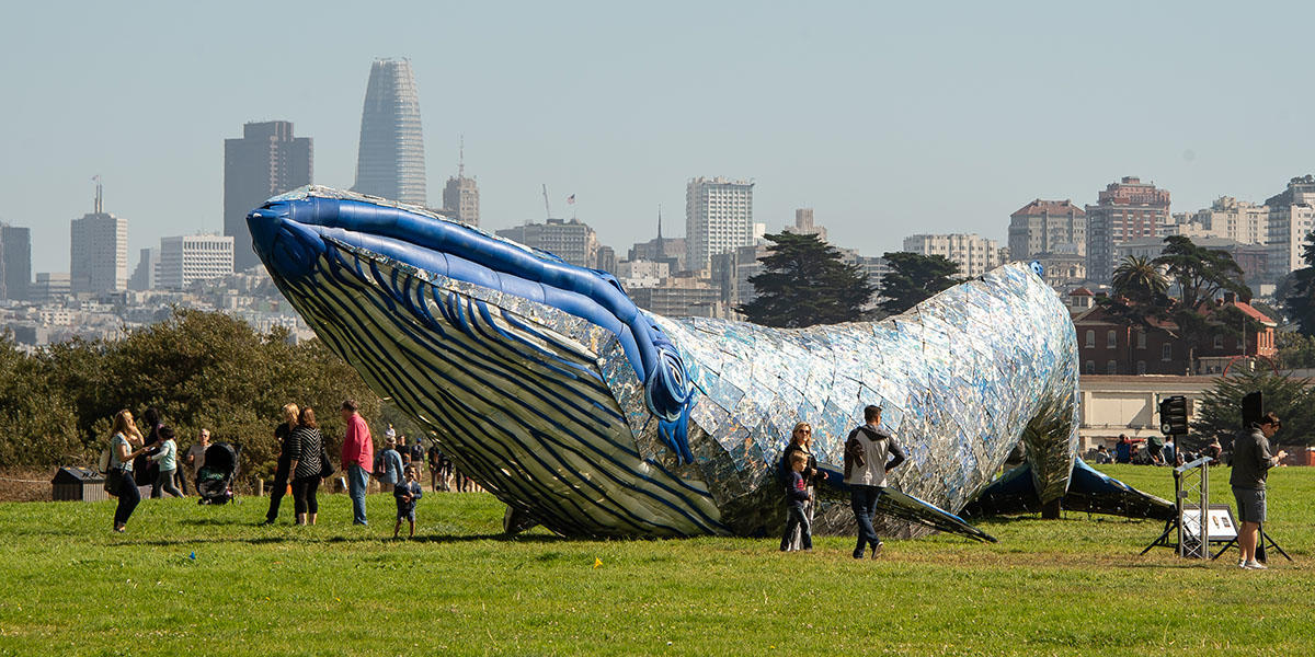 People wander around a huge sculpture of a blue whale, with the San Francisco skyline in the distance