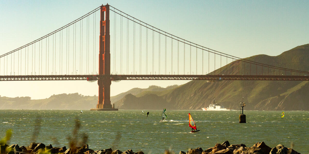Windsurfers cruise across bay waters with the Golden Gate Bridge spanned across the background.
