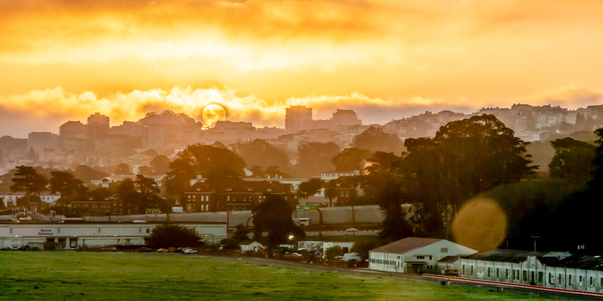 The bright yellow sun rises and shines through clouds over San Francisco skyline and Crissy Field.