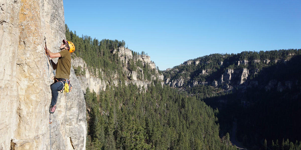 A person scales a rock wall outdoors overlooking a forest.