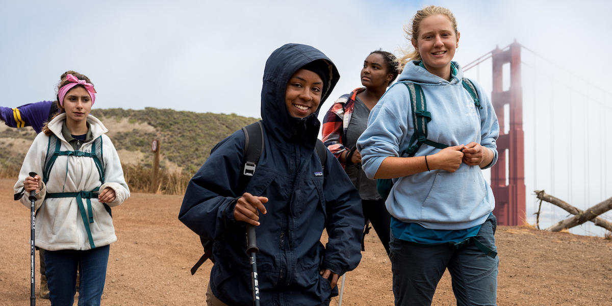 Four hikers trek down the SCA Trail in the Marin Headlands with the Golden Gate Bridge in the background.