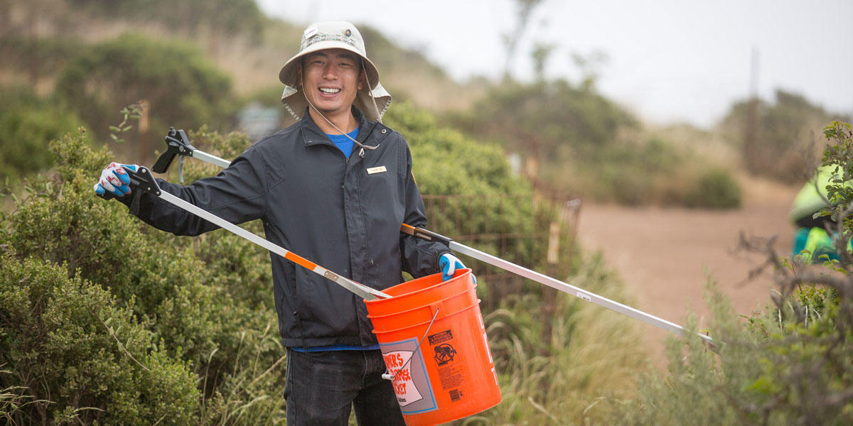 A volunteer holds a bucket and debris grabbing tool on a trail.