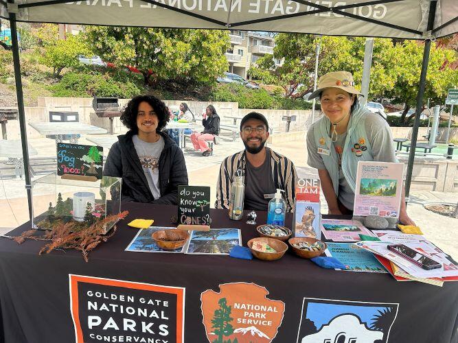 Three staff people pose under a tent with a black table covered in activities and green park background
