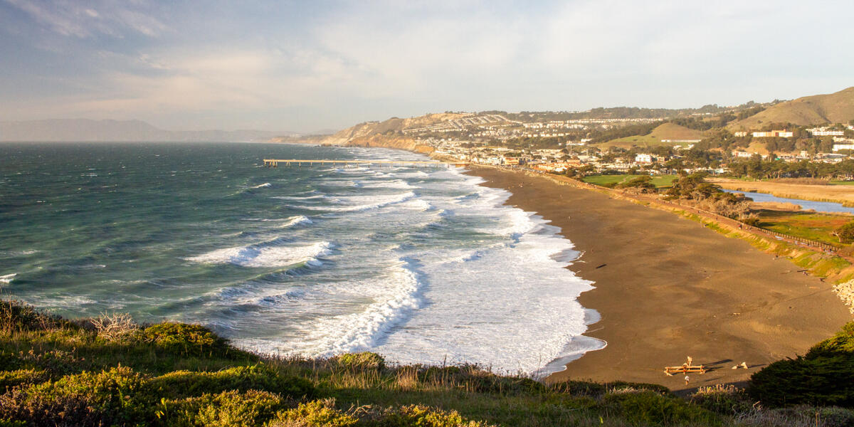 An expansive view of the coastline in Pacifica, CA from Mori Point. A long fishing pier stands out over the beach.