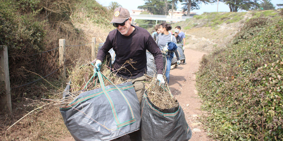 A volunteer carries large bags of vegetation while walking on a trail