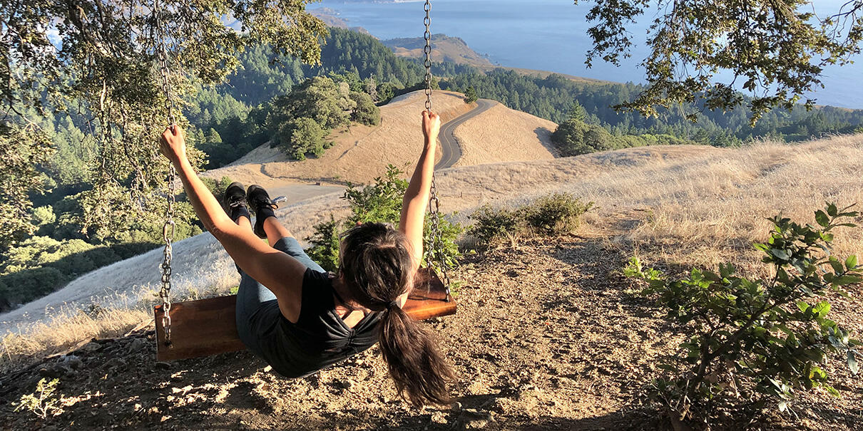 A person swings from a tree with a view of Mt. Tam and the Pacific Ocean.