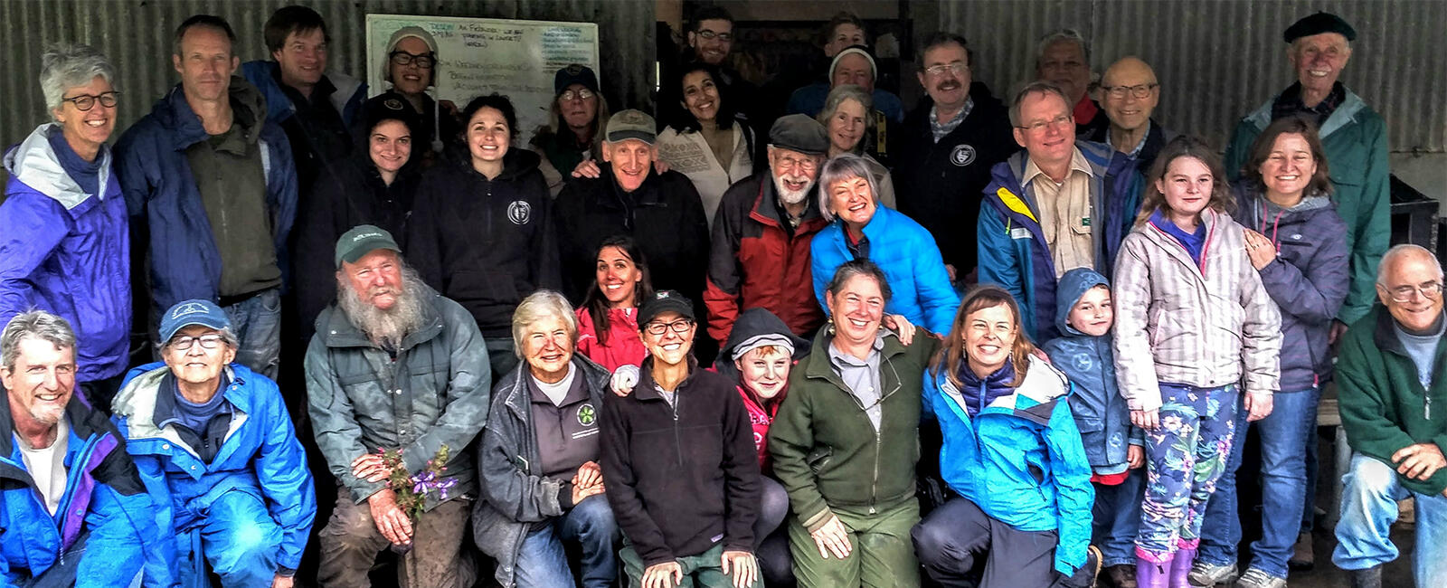 A large group smiles while posing for a photo in front of a metal barn.