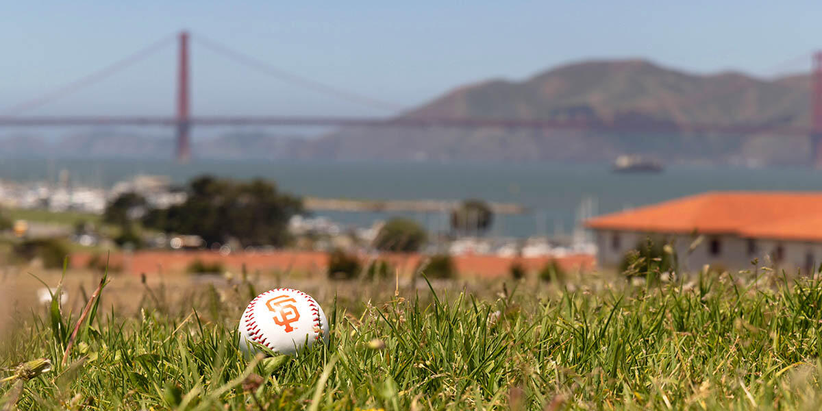 A baseball with the SF Giants logo lays on the green lawn at Fort Mason with a view of the Golden Gate Bridge in the background.