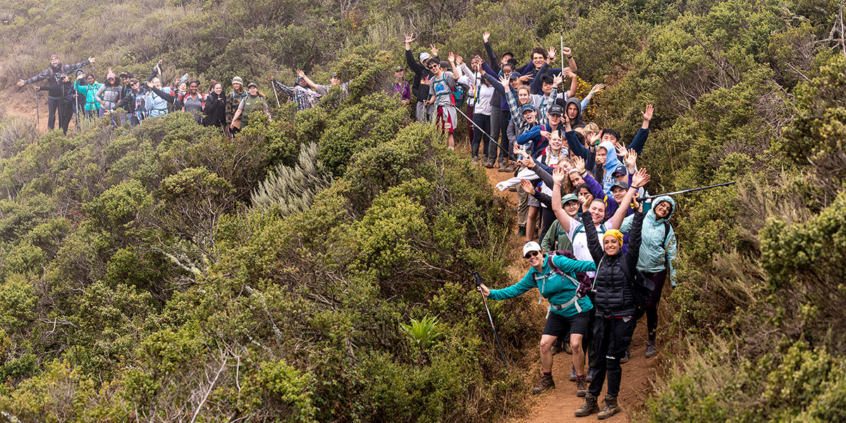 people standing on a trail waving at the camera with bushes in landscape