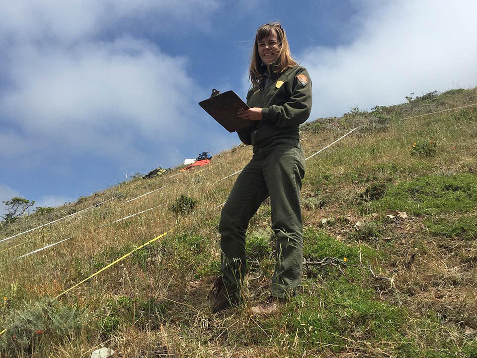 Alison Forrestel looks up for a smile while holding a clipboard among vegetation plots in a hilly grassland habitat.