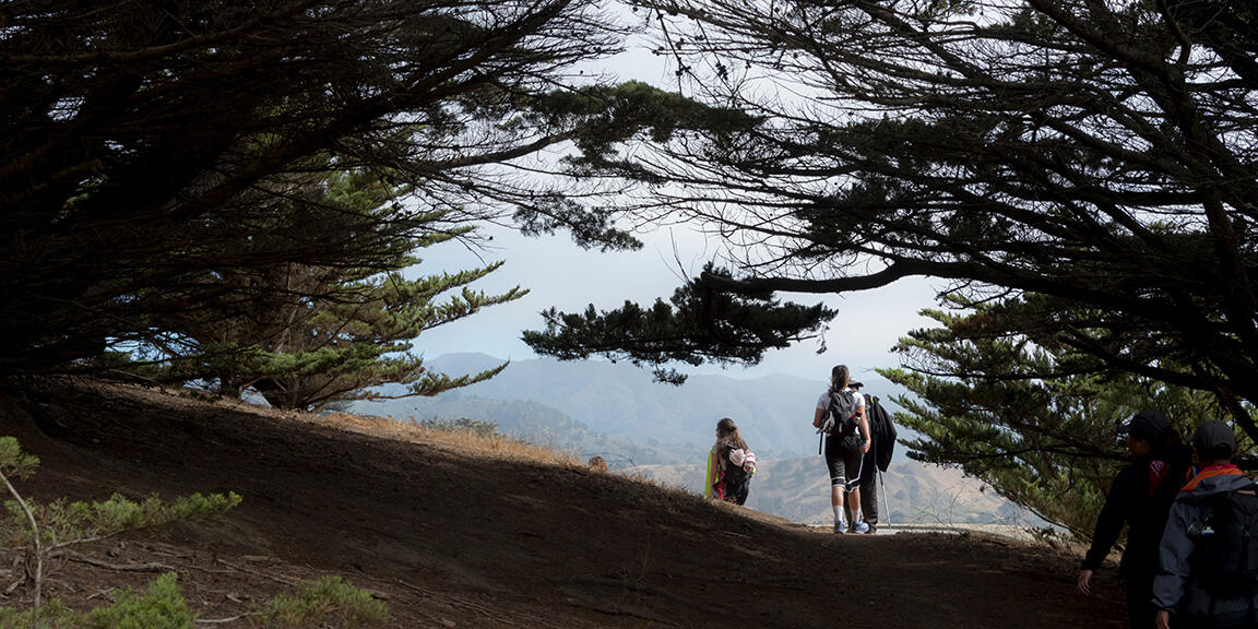 People hiking down a winding path through trees at Rancho Corral de Tierra, sloping hillsides are visible ahead and in the background.