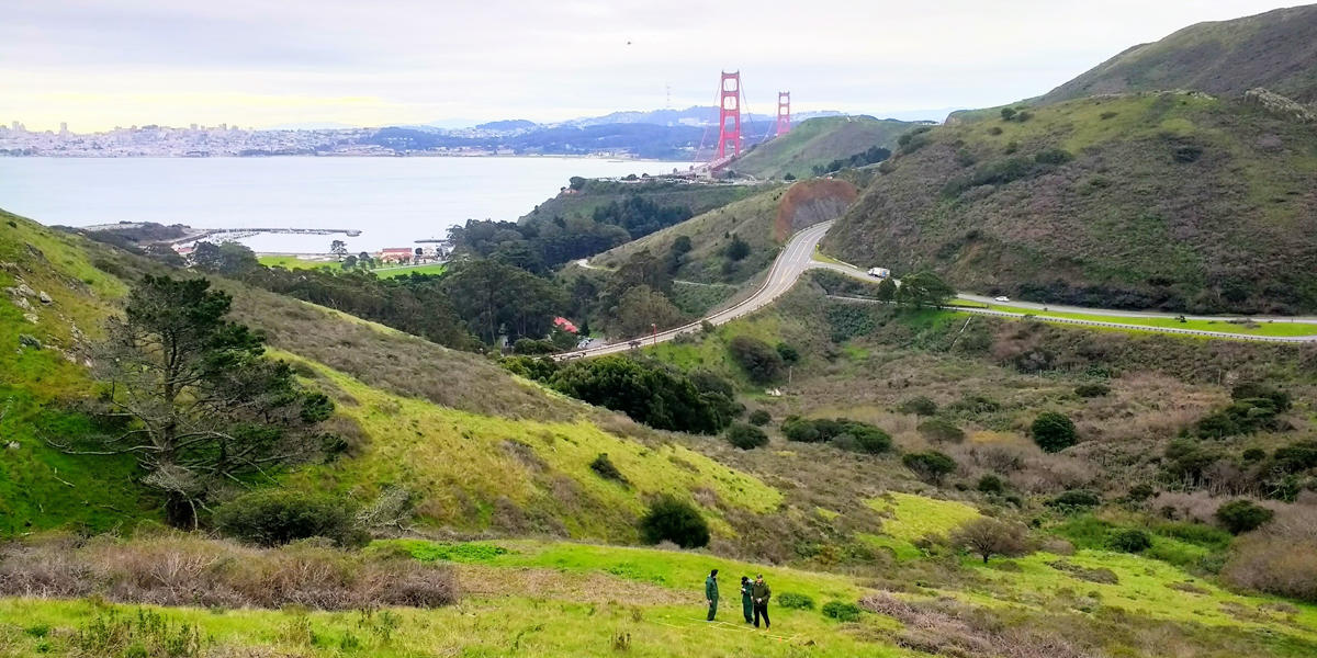 Three people stand on a hillside overlooking the Golden Gate Bridge