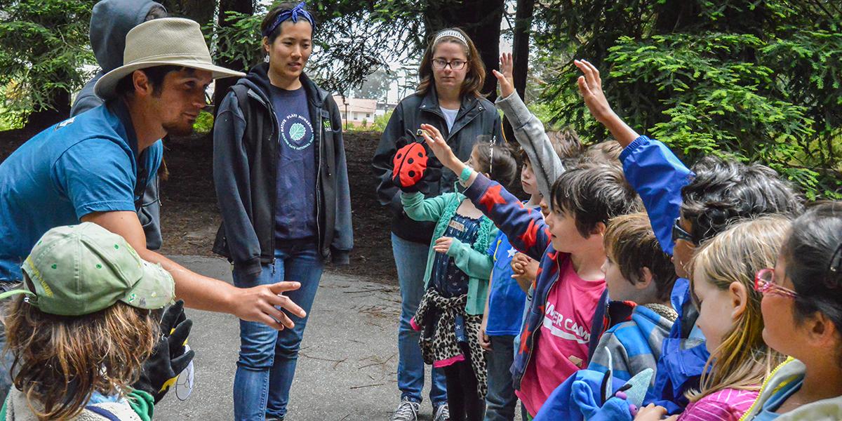 Presidio Nursery staff answer questions from Crissy Field Center Summer Campers. Presidio Nursery staff speaks with a group of young children with their hands raised for questions.