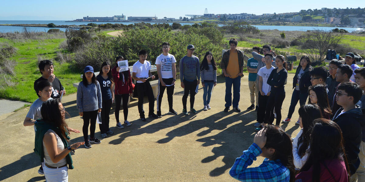 High school students stand in a circle outdoors while listening to an educator describe the natural environment surrounding them