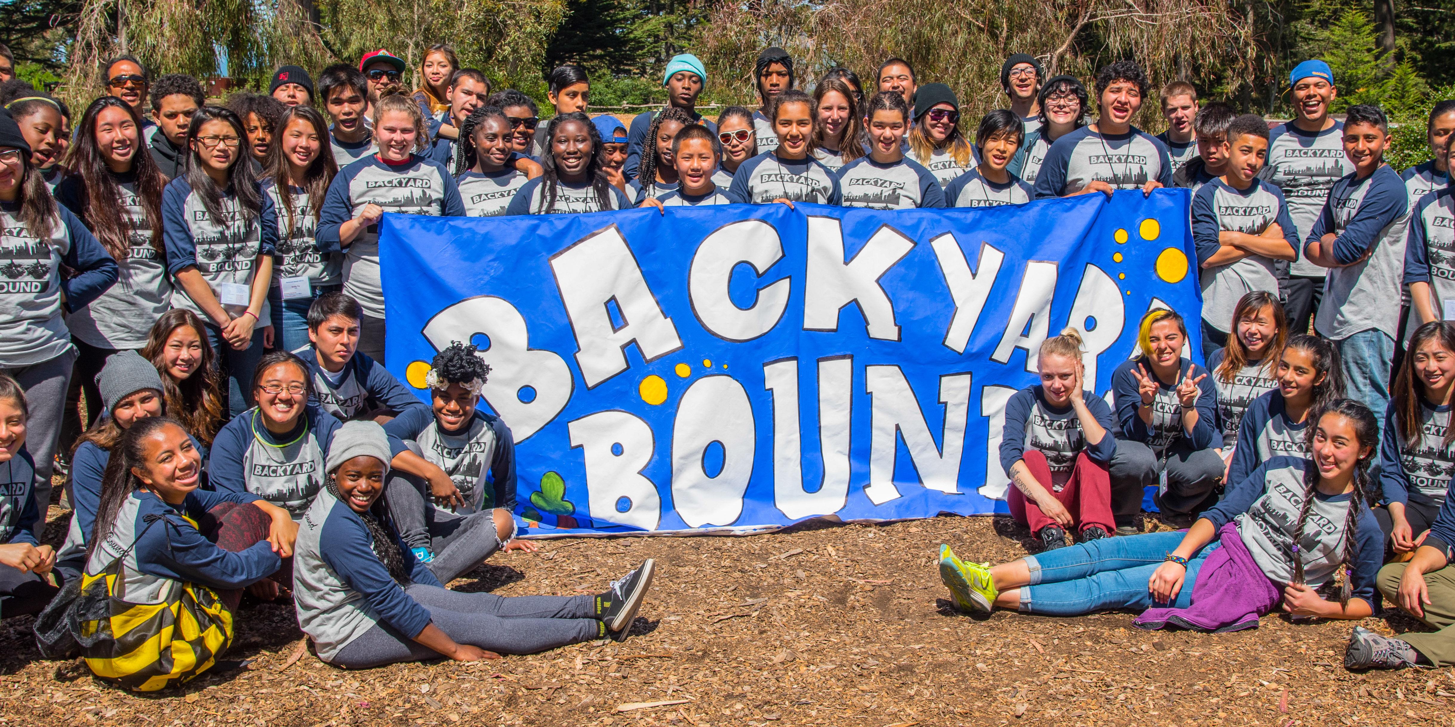 A large group of youth and youth workers gather around a banner reading Backyard Bound