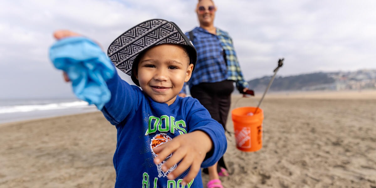 A happy child smiles while presenting a piece of collected trash to the camera during Coastal Clean Up Day at Ocean Beach