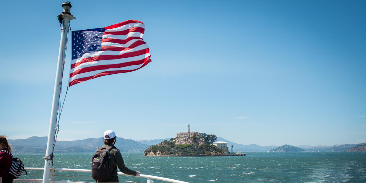 A park visitor looks toward Alcatraz Island from the ferry. An American flag waves in the breeze as they cruise over bay waters.