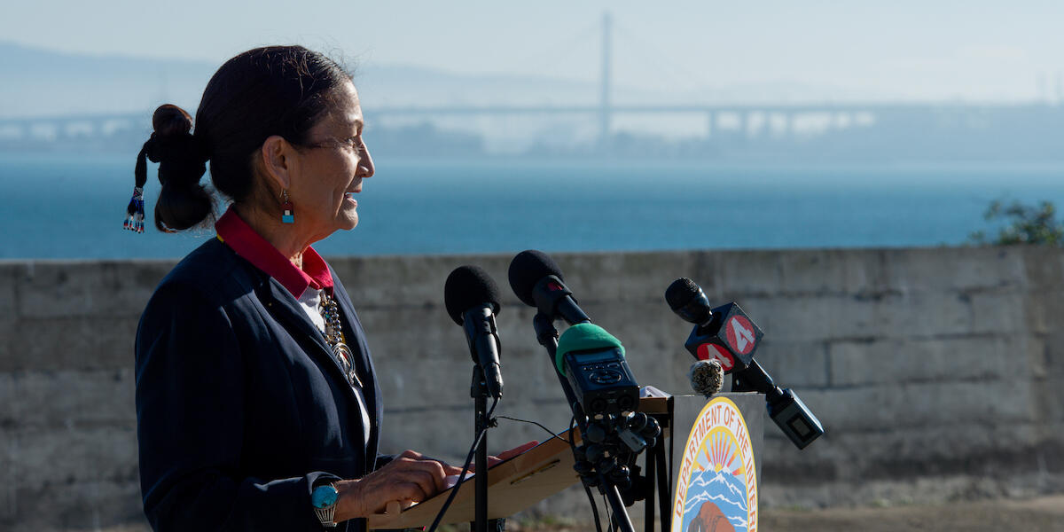 Secretary of the Interior, Deb Haaland, speaks at a press conference on Alcatraz Island with the Bay Bridge in the background.