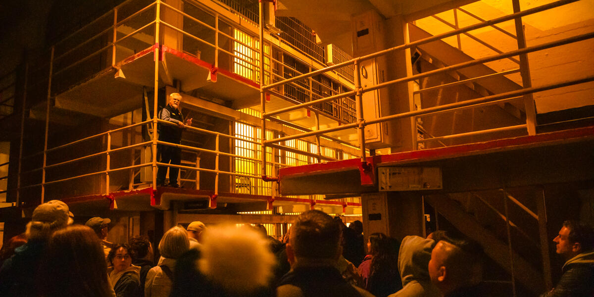 An Alcatraz tour-guide demonstrates the prison cell door mechanisms to a crowd under dim light during the night tour