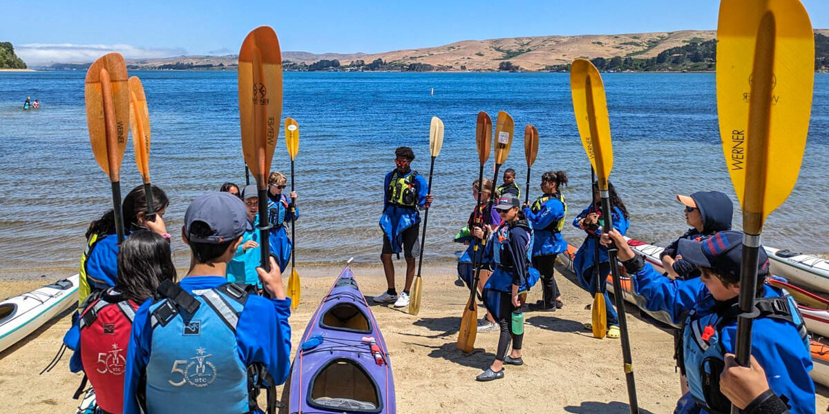 Crissy Field Center youth participants on the sunny shore of Hearts Desire Beach with kayaks and paddles