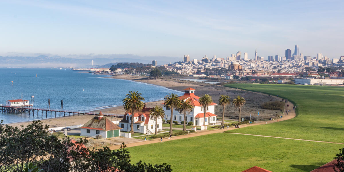 Aerial view of Crissy Field, buildings, the Warming Hut, palm trees, the golden gate, bay, and San Francisco city skyline.