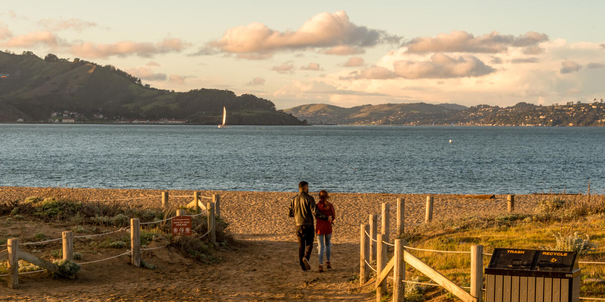 Visitors step out onto Crissy Field East Beach where the sunset casts warm light over the Golden Gate and San Francisco bay.