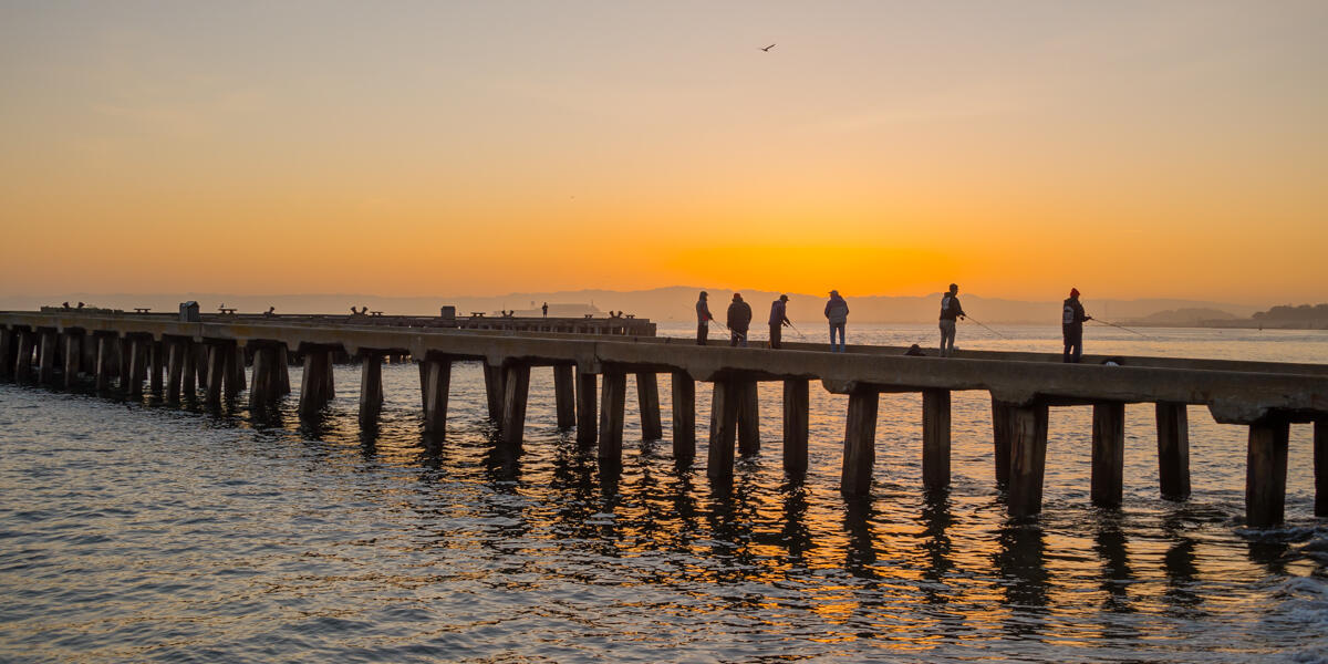 The sun rises over the bay waters, visitors are seen fishing off of the pier at Crissy Field.