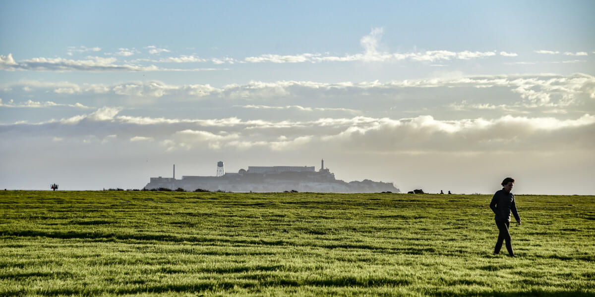 A view of Alcatraz from Crissy Field. A park visitor walks across the green grass under the blue sky.