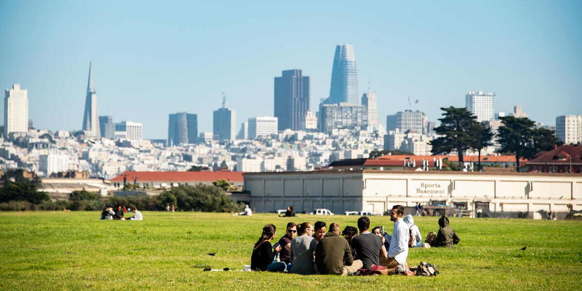Park visitors enjoy a picnic sitting on the lawn at Crissy Field amongst the San Francisco city skyline on a sunny day.