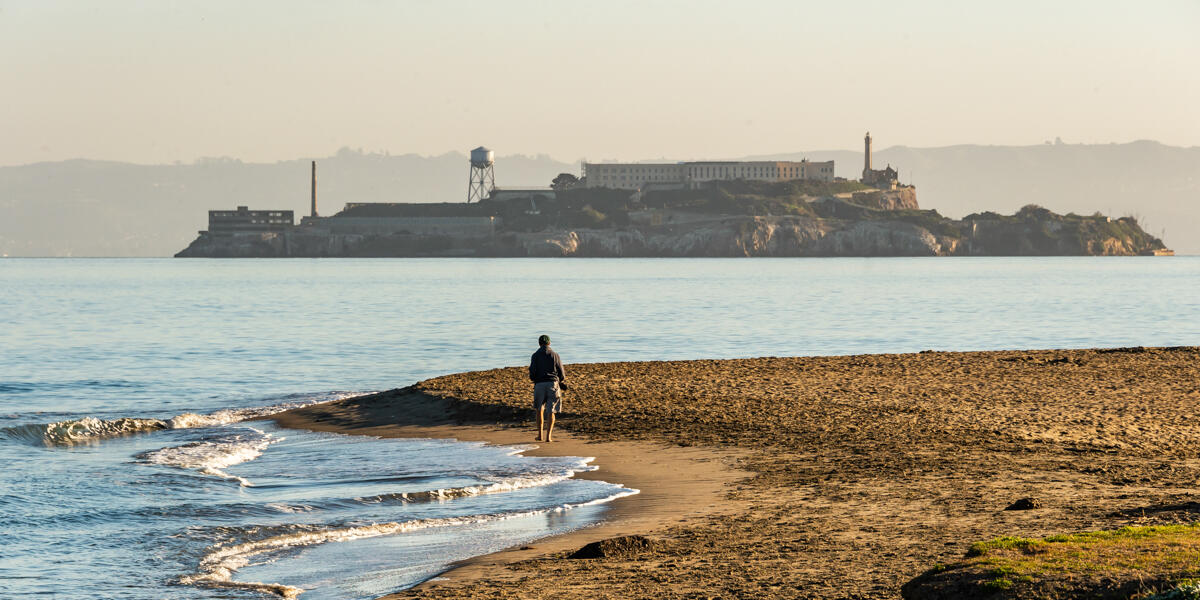 A park visitor walks along Crissy Field East Beach with a view of Alcatraz in the distance.