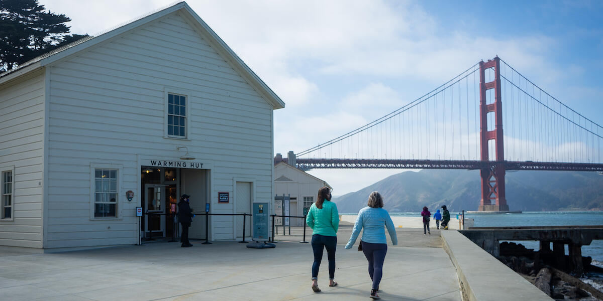 Two women walking down paved path towards the Golden Gate Bridge.