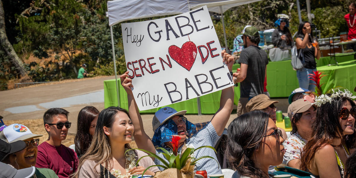 Guy wearing hat holding up sign reading, "Hey Gabe, serenade my babe!" at the Give Back Kick Back at Lands End in San Francisco.