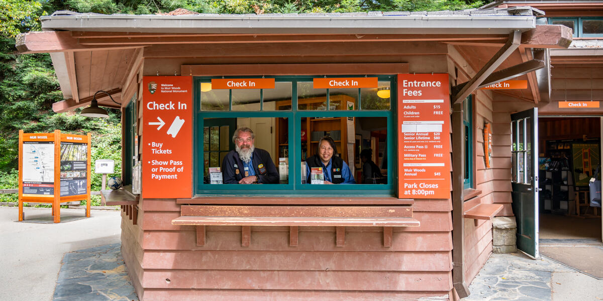 Two smiling Parks Conservancy staff members stationed in the Muir Woods check in kiosk windows.