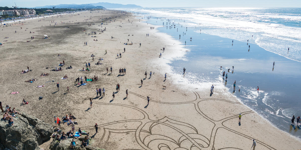 Aerial view of beach goers gathering around sand art at Ocean Beach.