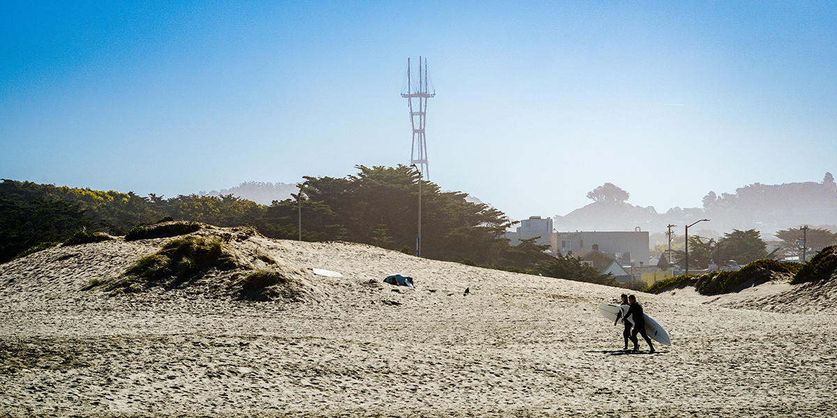 Surfers walk along Ocean Beach with Sutro Tower in the background