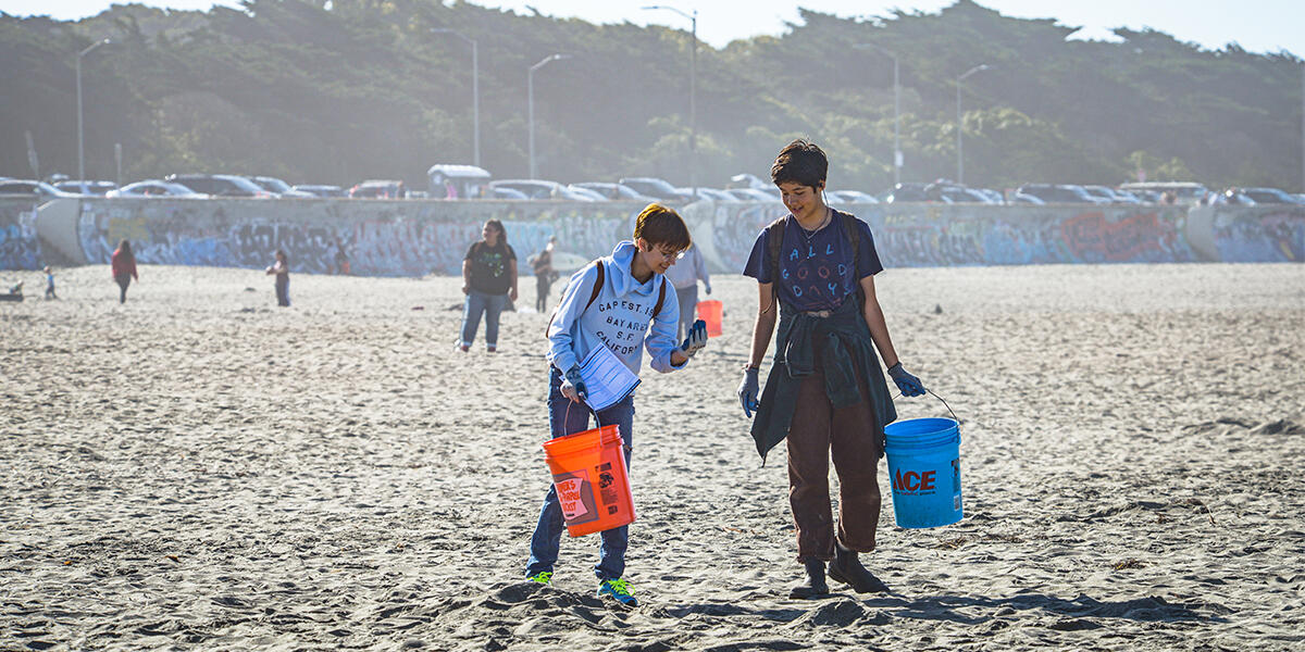 Two volunteers on trash pick up walk down Ocean Beach with buckets. 