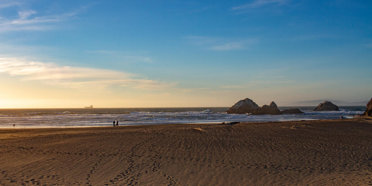 The open expanse of Ocean Beach shows an evening view of sandy shore, park visitors, ocean waves, and a commercial ship upon the horizon.