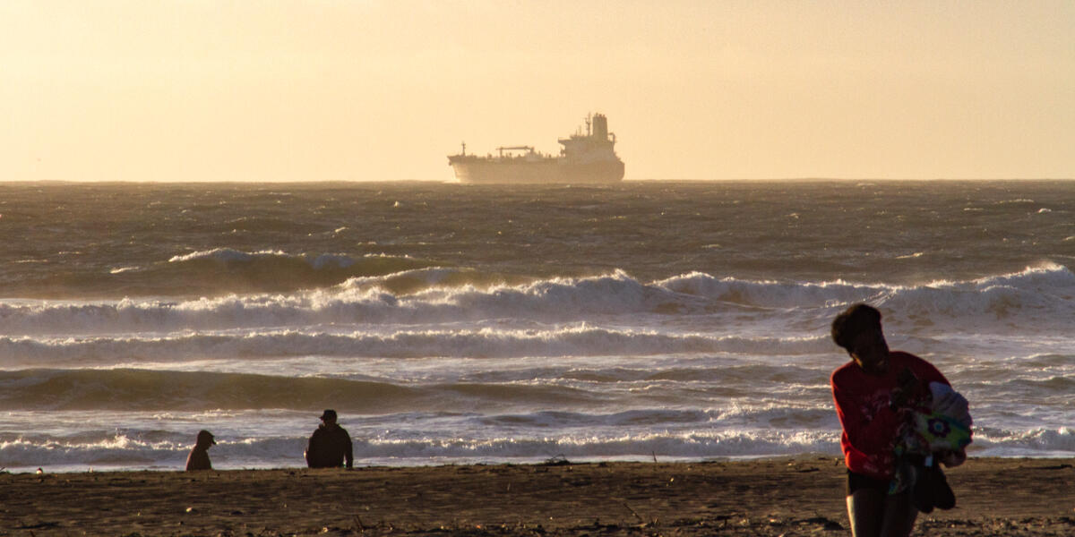 A large ship floats over the waves, along the horizon of Ocean Beach as park visitors enjoy the view of a sunset.