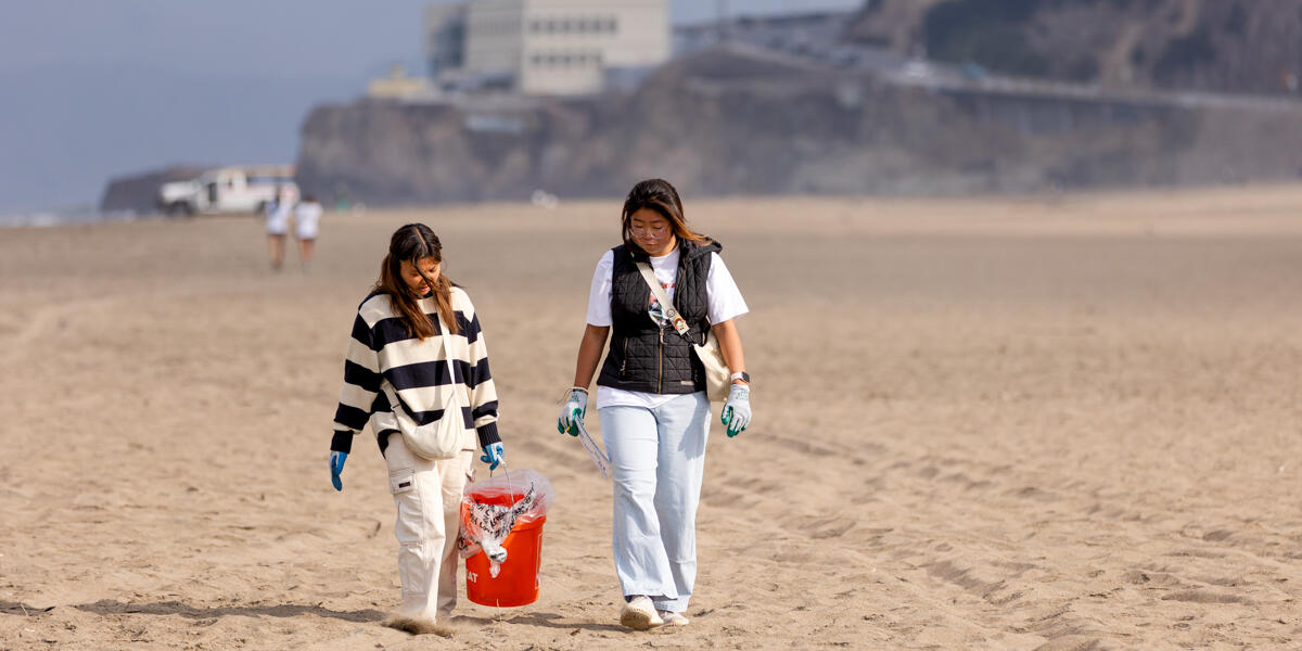 Two women carry bright orange buckets while volunteering for beach cleanup at Ocean Beach