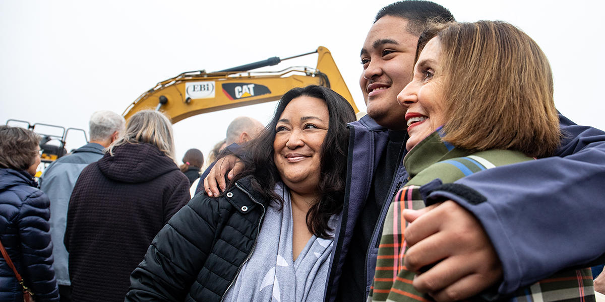 Three people embrace in front of excavator.