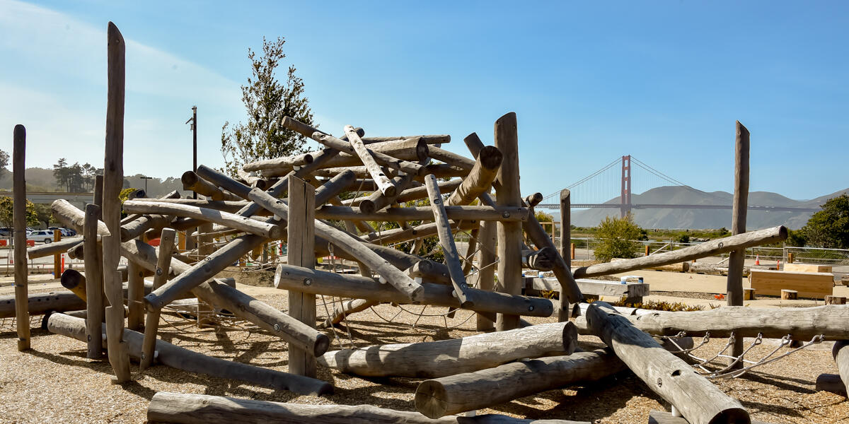 A playground structure made of wood logs at the Presidio Tunnel Tops Outpost, the Golden Gate Bridge is seen in the background.
