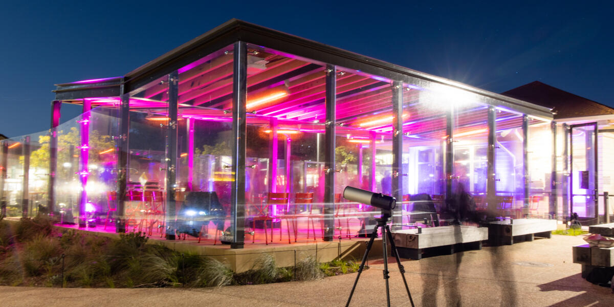 Long exposure photo of an enclosed outdoor picnic area in the Presidio glows with bright pink lights with an astronomy telescope seen in the foreground