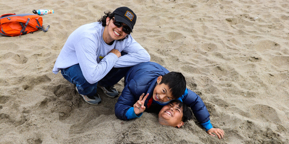Three smiling and laughing youths posing at Baker Beach, one is buried in the sand with their head revealed