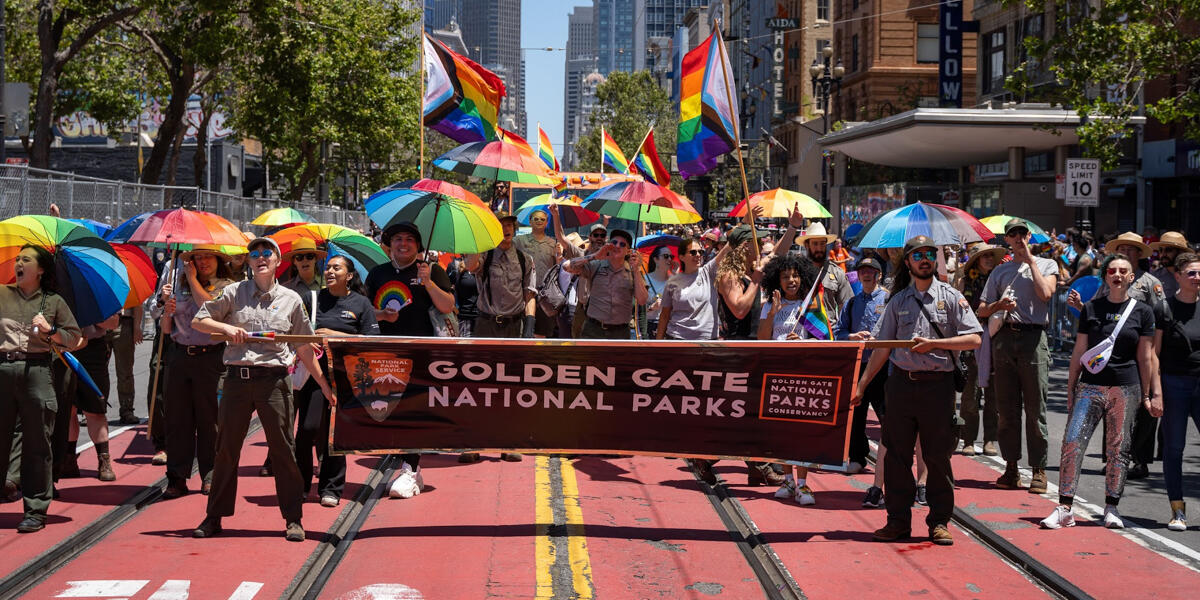 Smiling Parks Conservancy staff and NPS Rangers hold a Golden Gate National Parks banner and rainbow umbrellas in the Pride Parade.